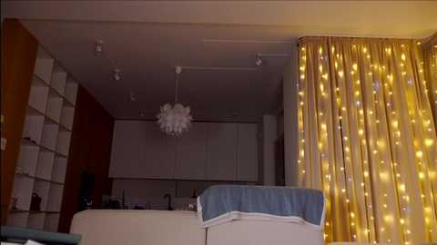 Media: Video of a modern, dimly lit living room with white furniture, a yellow string-lit curtain, a blue blanket, and a white chandelier.