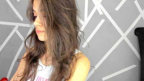 Media: Video of a young woman with long, wavy brown hair, wearing a white tank top, against a geometric, grey and white patterned wall.