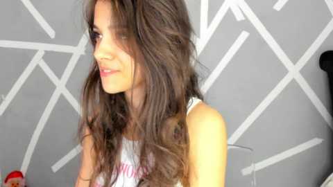 Media: Video of a young woman with long, wavy brown hair, wearing a white tank top, smiling softly. She is indoors, with a geometric white patterned wall in the background.