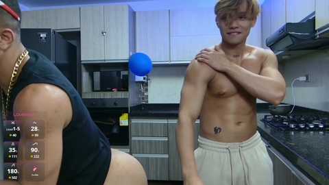 Media: Video of a shirtless, muscular, Asian man with a tattoo on his chest, wearing beige shorts, standing in a modern kitchen with cabinets and appliances.