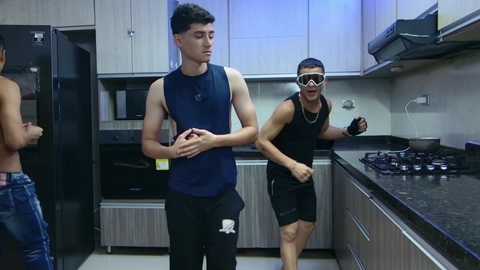 Media: Video of three shirtless young men in a modern kitchen, two wearing black sleeveless shirts, one in a blue one, playing with a football, and another man in black shorts and a mask, standing near a stove.