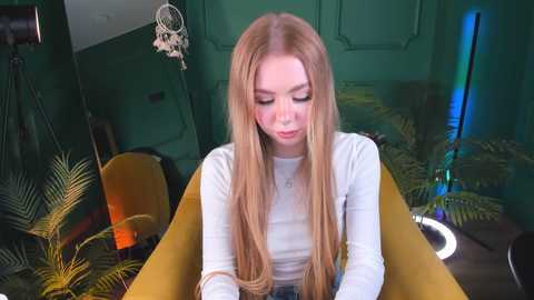 Media: Video of a blonde, fair-skinned woman with long hair, wearing a white top, sitting in a mustard-yellow chair, surrounded by potted plants in a dimly lit, green-walled room.