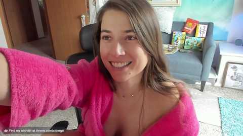 Media: Video of a young woman with long brown hair, smiling, wearing a bright pink robe, seated on a gray couch in a cozy living room.