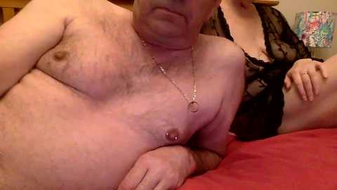 Media: A video showing a topless, middle-aged man with a light complexion and a silver necklace, lying on a red bed next to a partially visible woman in black lace lingerie.