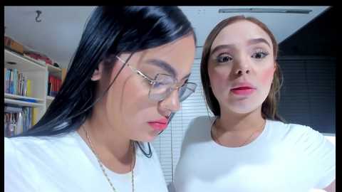 Media: Video of two young women with light skin, one with straight black hair, glasses, and a white top, the other with light brown hair and a white top, both indoors.