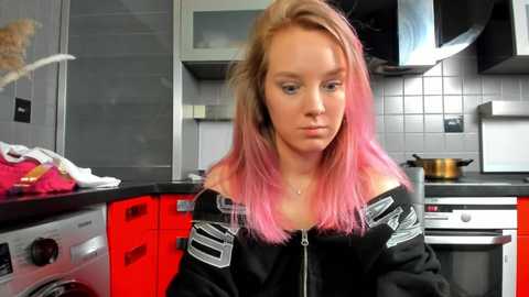 Media: Video of a young woman with long, pink and blonde hair, wearing a black off-shoulder hoodie, sitting in a modern kitchen with red and grey cabinets, silver appliances, and a copper pot on the stove.