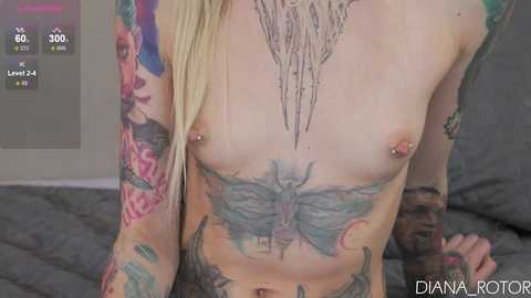 Media: Video of a topless, pale-skinned person with blonde hair, showcasing extensive tattoos including a large butterfly on the chest, colorful floral designs on the arms, and a geometric design on the neck. The background features a gray bed.