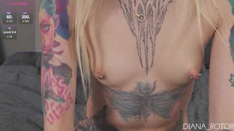 Media: Video of a topless, fair-skinned woman with blonde hair, showcasing detailed tattoos including floral and butterfly designs on her chest, with nipples pierced. The background features a bed with gray sheets.