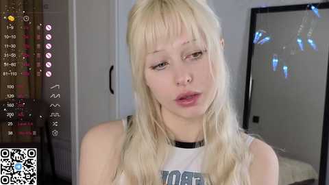 Media: Video of a fair-skinned, blonde-haired woman with blunt bangs, wearing a white tank top, standing indoors. Background includes a mirror, curtains, and digital camera interface.