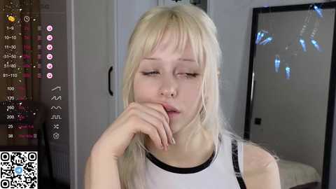 Media: A video of a young, fair-skinned, blonde woman with long hair, wearing a white tank top, resting her chin on her hand, looking tired. The background shows a dimly lit room with a bed and a mirror.