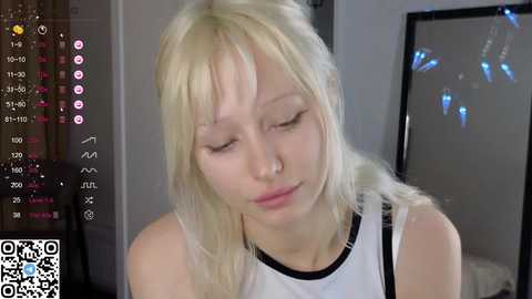 Media: Video of a young, pale-skinned, platinum blonde woman with long hair, wearing a white tank top, eyes closed, in a dimly lit room with a mirror and digital camera interface.