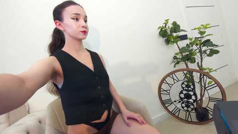 Media: A video of a young woman with light skin and brown hair in a ponytail, wearing a black sleeveless vest and black panties, sitting on a beige chair in a minimalist room with a large, ornate clock and a potted plant.