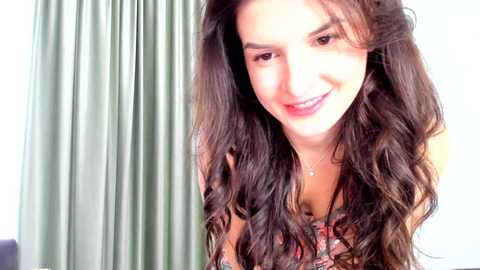 Media: Video of a young woman with long, wavy brown hair and fair skin, smiling. She wears a floral-patterned top. Green curtains and white walls form the background.