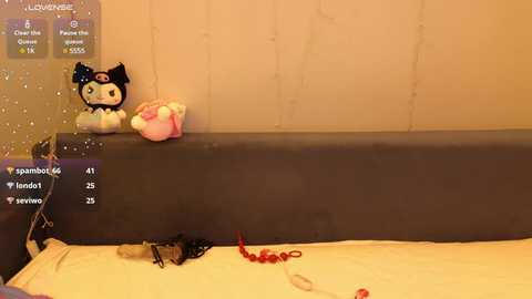 Media: A video of a bedroom with a dark gray headboard, featuring a plush black cat toy with a red bow and a pink bunny toy on the bed. The background has beige curtains and a digital display showing user details.