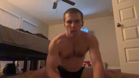 Media: Video of a shirtless, muscular man with short hair wearing black briefs, kneeling on a carpeted floor in a dimly lit bedroom with a bed, window, and closed door.