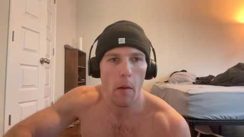 Media: Video of a shirtless, fair-skinned man with a black beanie, large headphones, and a tongue sticking out. He's in a dimly-lit bedroom with a bed and white door in the background.