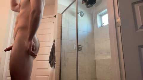 Media: A video of a nude, fit, light-skinned man standing in a modern, tiled bathroom with a shower, towels, and a window. He has an erect penis.