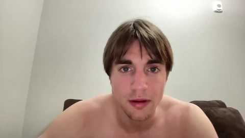 Media: Video of a young, shirtless Caucasian man with short brown hair, fair skin, and a neutral expression, sitting on a dark brown couch against a plain white wall.