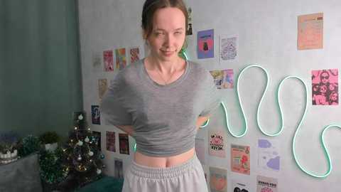 Media: Video of a young woman with fair skin, light brown hair tied back, wearing a grey crop top and grey pants, standing in a colorful room adorned with neon signs and framed artwork.