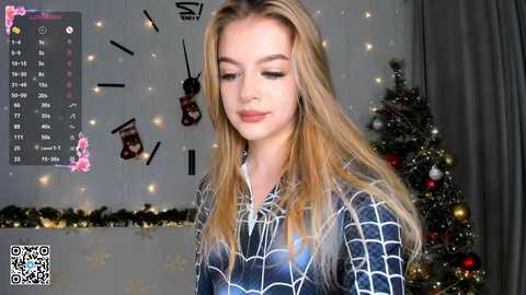 Media: Video of a young woman with long blonde hair, wearing a blue Spider-Man shirt, standing next to a decorated Christmas tree, with a calendar and holiday lights in the background.