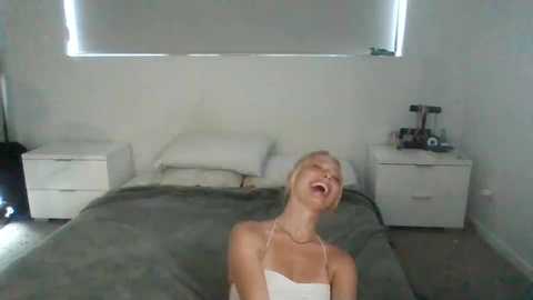 Media: Video of a blonde woman in a white dress, screaming with eyes closed, in a minimalist bedroom with white walls, a bed, and two white nightstands.