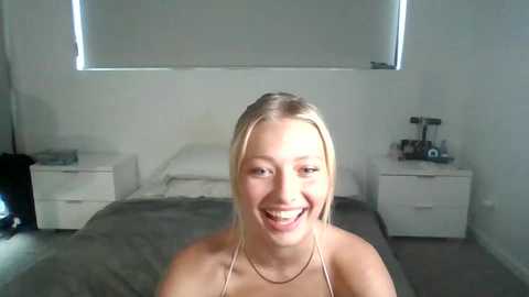 Media: Video of a smiling young woman with blonde hair, light skin, and a small build, standing in a minimalist bedroom with white walls, a bed, and white furniture.