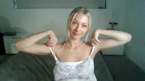 Media: Video of a young blonde woman with fair skin, small breasts, and slender physique, wearing a white lace camisole, posing confidently with arms raised in a bedroom with a white bed and nightstand.