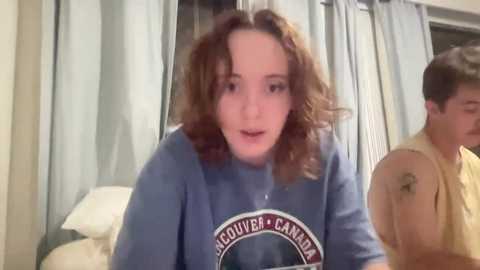 Media: Video of a young, light-skinned woman with curly brown hair, wearing a blue \"Saginaw Chippewas\" shirt, and a man in a yellow shirt, both indoors.