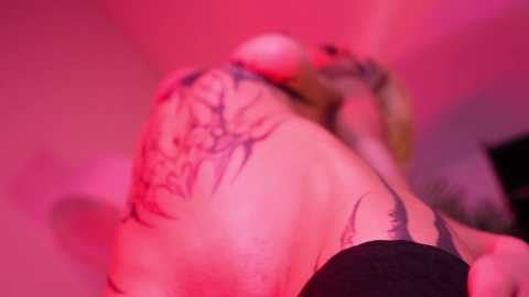 Video of a tattooed person's back in a dimly lit, pink-hued room, featuring intricate, black tribal designs. The subject's shoulder and upper arm are visible, with a blurred background suggesting an intimate, possibly private setting.