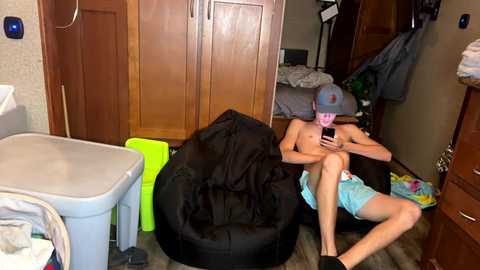 Media: Video of a shirtless man with a mask, blue shorts, sitting on a black bean bag chair in a cluttered bedroom, surrounded by laundry and wooden furniture.