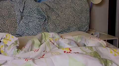 Media: Video of a messy bed with a white comforter featuring green, red, and yellow patterns, and a black-and-white spotted pillow. The room's background shows a beige wall with a small, round white lamp.