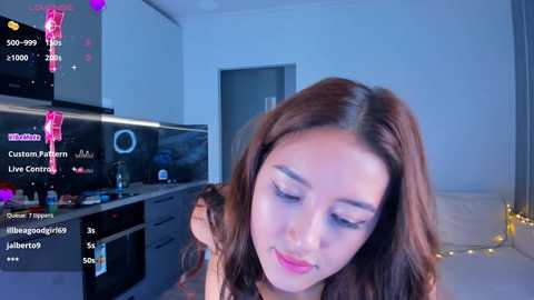 Media: Video of an Asian woman with long brown hair, light skin, and closed eyes, sitting in a modern, well-lit room with a flat-screen TV, kitchen counter, and string lights in the background.