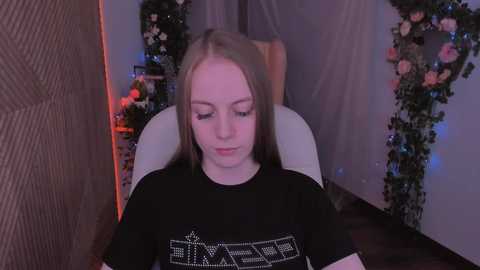 Media: Video of a young, fair-skinned woman with long blonde hair, wearing a black T-shirt with a white graphic, seated in a chair in a dimly lit room with floral decorations.