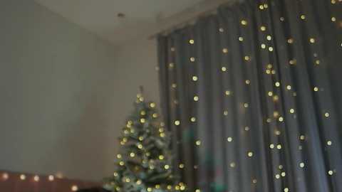 A video of a softly lit room featuring a Christmas tree decorated with white lights and a blue-gray curtain adorned with golden string lights.