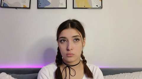 Media: Video of a young woman with light skin, brown hair in two braids, wearing a white top and black choker, against a white wall with abstract art.