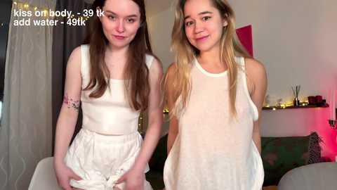 Media: Video of two young women in white, sheer tops and shorts, standing indoors. One has long brown hair, the other blonde. Background features a green couch and dimly lit room.