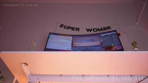 Media: A video of a desk with two computer monitors displaying various screens. A banner with \"SUPER WOMAN\" written in black letters hangs on the wall behind the monitors. The background is a plain, light-colored wall.