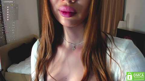 Media: Video of a fair-skinned woman with long, straight auburn hair, wearing a white cardigan and silver necklace. Her lips are slightly parted, showing full, pink lips. Background includes a bed with a white comforter and a wooden nightstand.