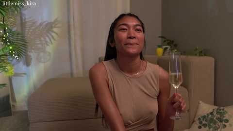 Media: Video of an East Asian woman with straight, dark hair, wearing a beige sleeveless top, holding a glass of white wine, seated in a modern living room with beige sofa, green plants, and soft lighting.