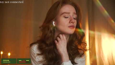 Media: Video of a fair-skinned woman with long, wavy brown hair, wearing white earrings, eyes closed in a contemplative pose, in a warm, blurred background with golden light.