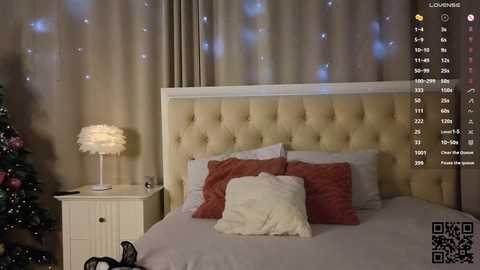 Video of a cozy, modern bedroom with beige tufted headboard, white walls, fluffy pillows, and a small white nightstand with a round lamp. A Christmas tree with ornaments is partially visible on the left.