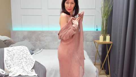 Media: Video of an Asian woman with medium-length black hair, wearing a sheer mauve robe, standing in a modern bedroom with gray curtains, white bed, and wooden side table.