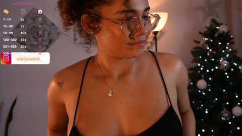 Media: Video of a young woman with curly hair, glasses, and a black tank top, standing near a Christmas tree in a cozy, warmly-lit room.