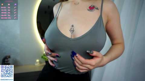 Media: Video of a woman with fair skin, large breasts, wearing a tight, low-cut gray tank top, revealing tattoos on her chest. She has long, dark hair. Background includes a round mirror and white curtains.