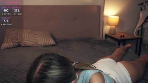 Media: Video of a woman lying on a bed, wearing a white tank top and shorts, with a bedside lamp casting a warm glow.