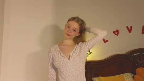 Media: Video of a young Caucasian girl with blonde hair, wearing a white floral-patterned long-sleeve top, standing in a bedroom with a wooden headboard and red heart decals on the wall.