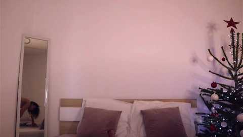 A video of a minimalist bedroom with a white wall, a bed with white pillows, a tall mirror, and a decorated Christmas tree in the corner.