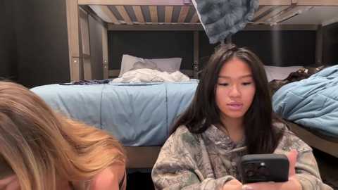 Media: Video of a young Asian woman with long black hair in a camo hoodie, sitting on a bunk bed in a dorm room. Her face is neutral, looking down at her phone. The room is cluttered with blue bedding and clothes.