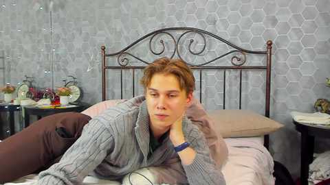 Media: Video of a young man with short, light brown hair, wearing a gray sweater, lying on a bed with gray, hexagonal-patterned wallpaper, clock, and potted plants in the background.