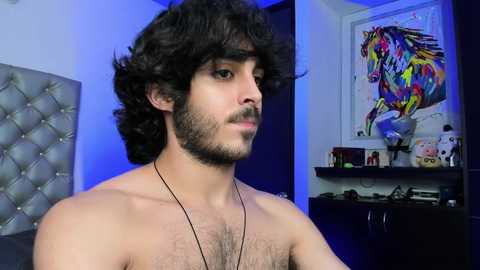 Media: Video of a shirtless man with curly dark hair and a beard, wearing a necklace. He stands in a room with a tufted gray headboard, colorful abstract painting, and a cluttered shelf.